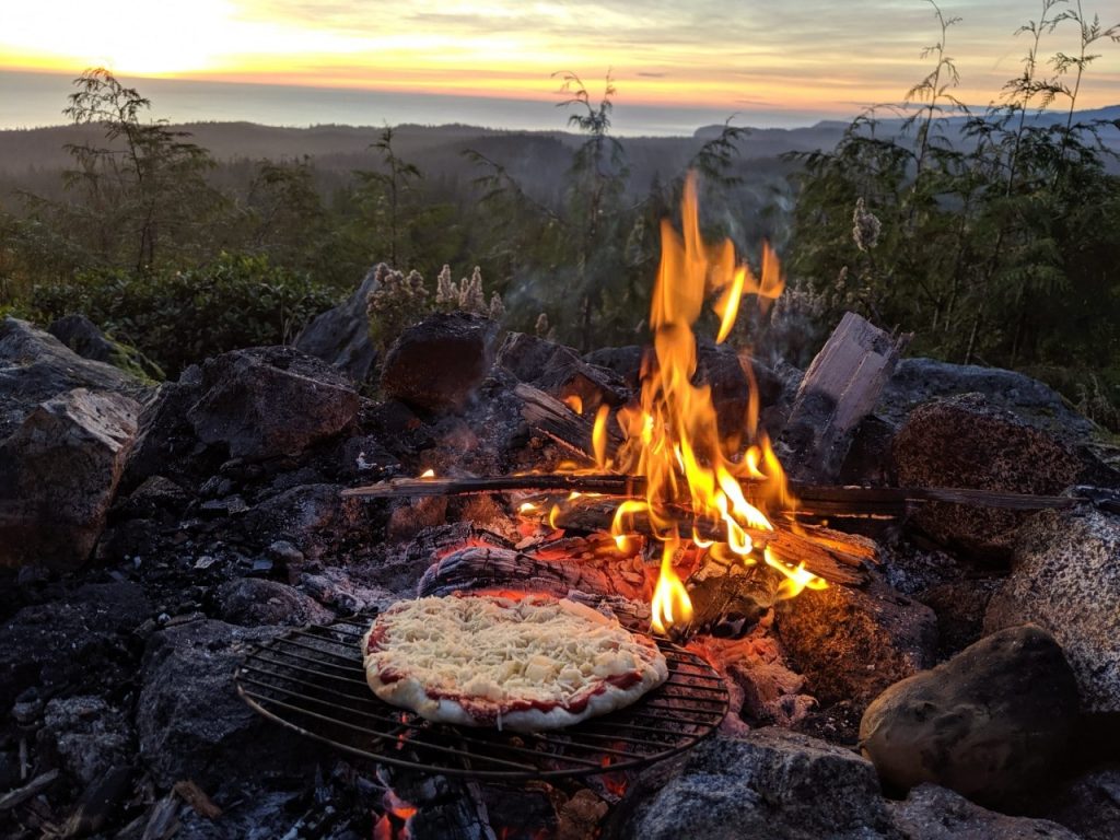 Homemade tomato and cheese pizza set on grill next to campfire with forest and ocean backgound