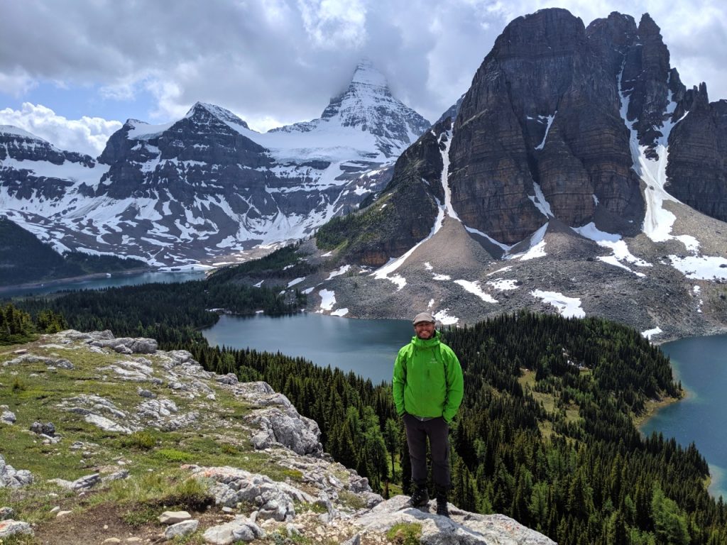 JR standing and looking at camera, with backdrop of alpine lakes and snow capped mountains in Mount Assiniboine Provincial Park, British Columbia, Canada