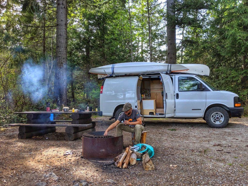 JR sitting at campfire in front of white van with open door, while camping in British Columbia