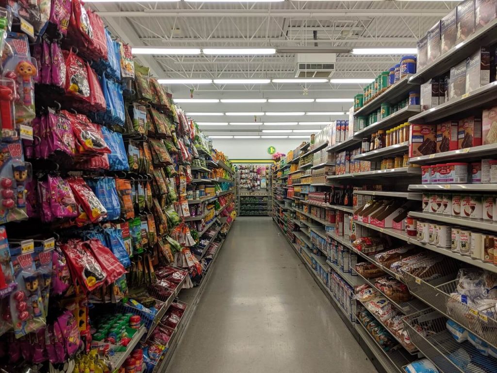 Looking down an aisle at a dollar store, with food on shelves on each side, with view of high ceiling above