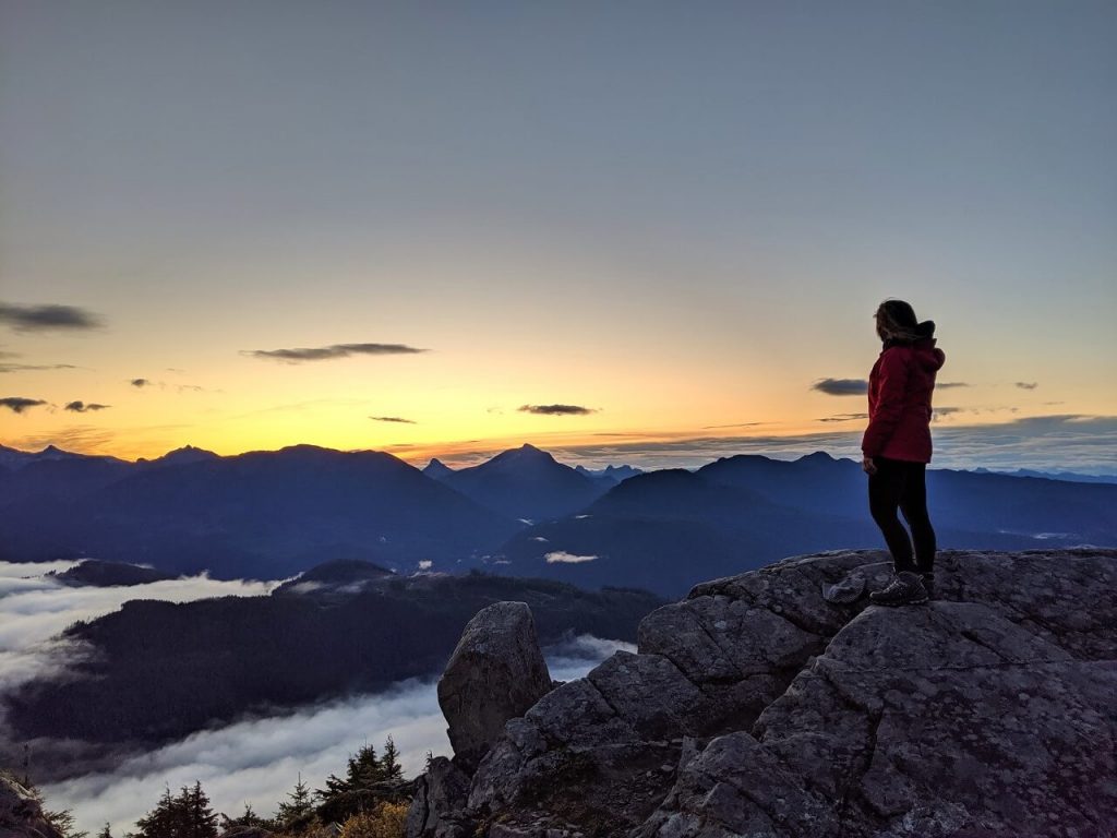 Gemma is standing on a rock on the right hand side of the photo, looking at the sunrise behind mountains below